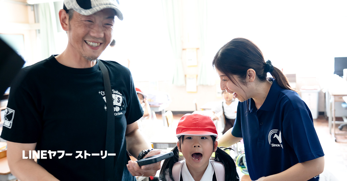 室内の明るい部屋で、赤い帽子をかぶった小さな子どもが口を大きく開けて前を向いている様子を、左右にいる大人2人が笑顔で見守っている写真。画像左下には「LINEヤフーストーリー」という白い文字が配置されている。