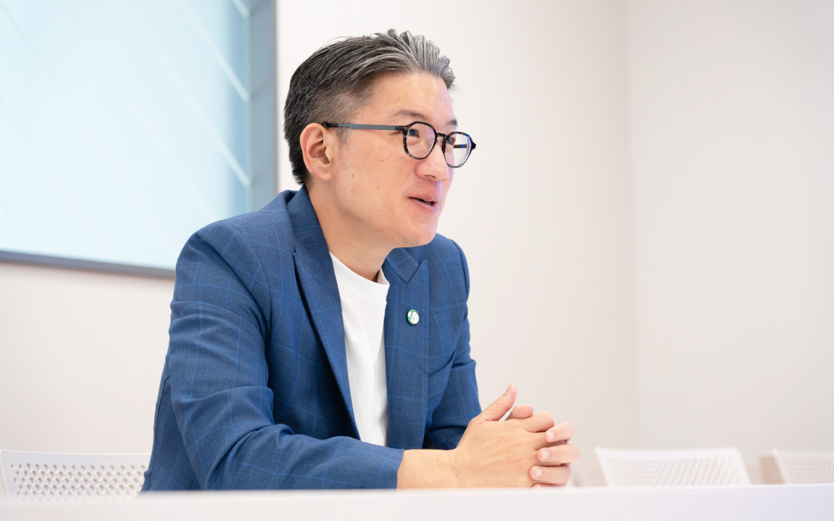 Yod Chinsupakulさんのバストショットである。グレーがかった黒髪を後ろに整え、黒縁の丸眼鏡をかけている。両手を軽く組みながら前傾姿勢で話している。横からのアングルで、口元にわずかに笑みを浮かべ、対話している様子である。