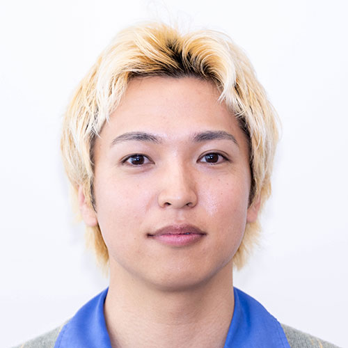 グレー背景で正面を向く男性、増田のバストショット。明るい金髪の短髪で、青いシャツとグレーのカーディガンを着用し、穏やかな表情を浮かべている。