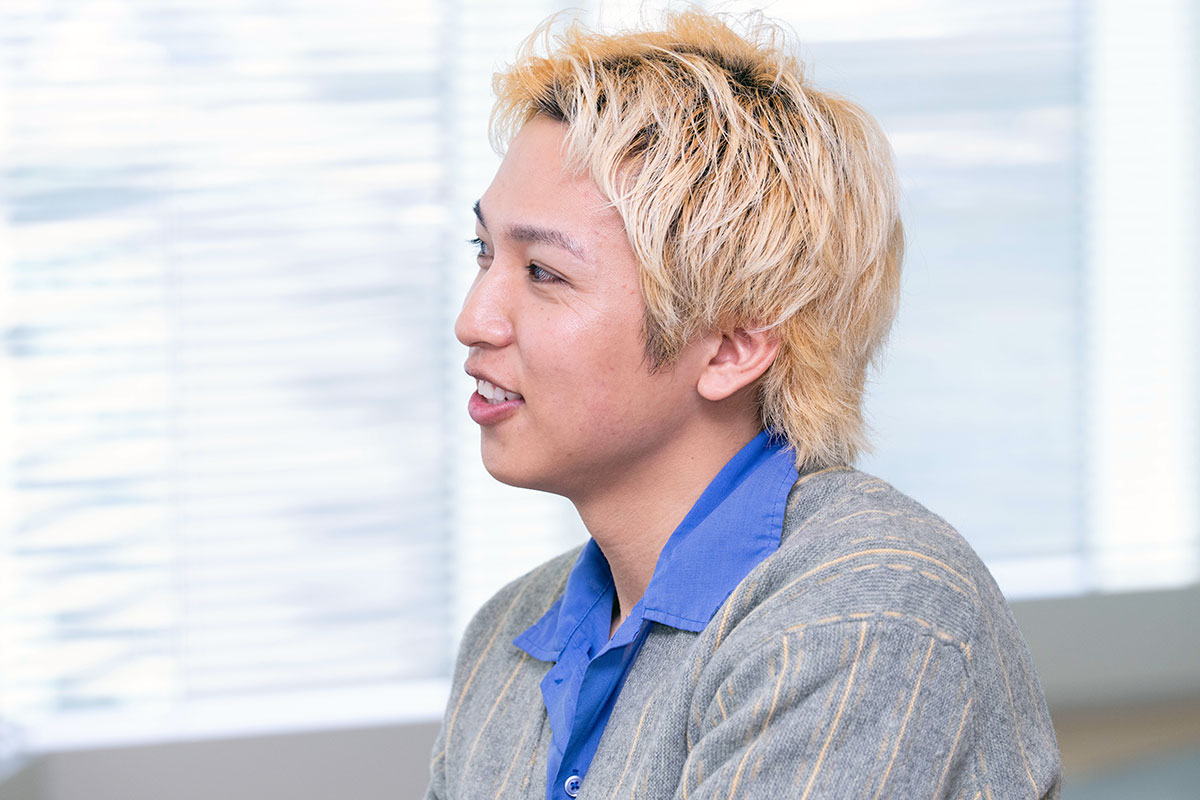 横向きで話す金髪の短髪男性、増田の写真。青いシャツとグレーのカーディガン姿で、穏やかな表情を浮かべながら会話している。背景にブラインドが見える。