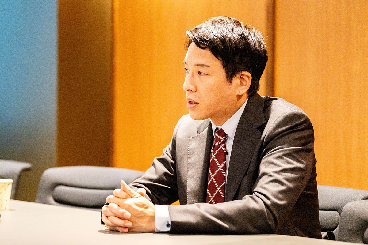 会議室のテーブルに座る金剛洙さんの写真。スーツ姿で両手を組み、横を向きながら話している