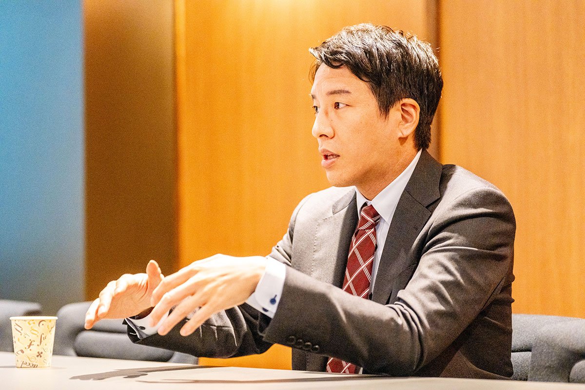 会議室で金剛洙さんが両手を広げながら話している写真。スーツ姿で横を向き、説明するように身振りを交えている