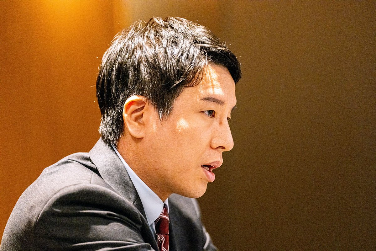スーツ姿の金剛洙さんを横から捉えた上半身写真。会議室で前方を見ながら話している