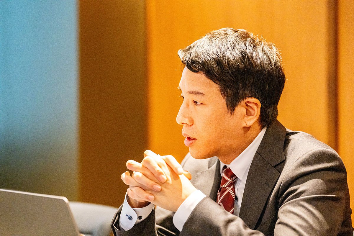 会議室で金剛洙さんが両手を組みながら話している写真。手前にはノートパソコンがあり、横を向いて落ち着いた表情を見せている