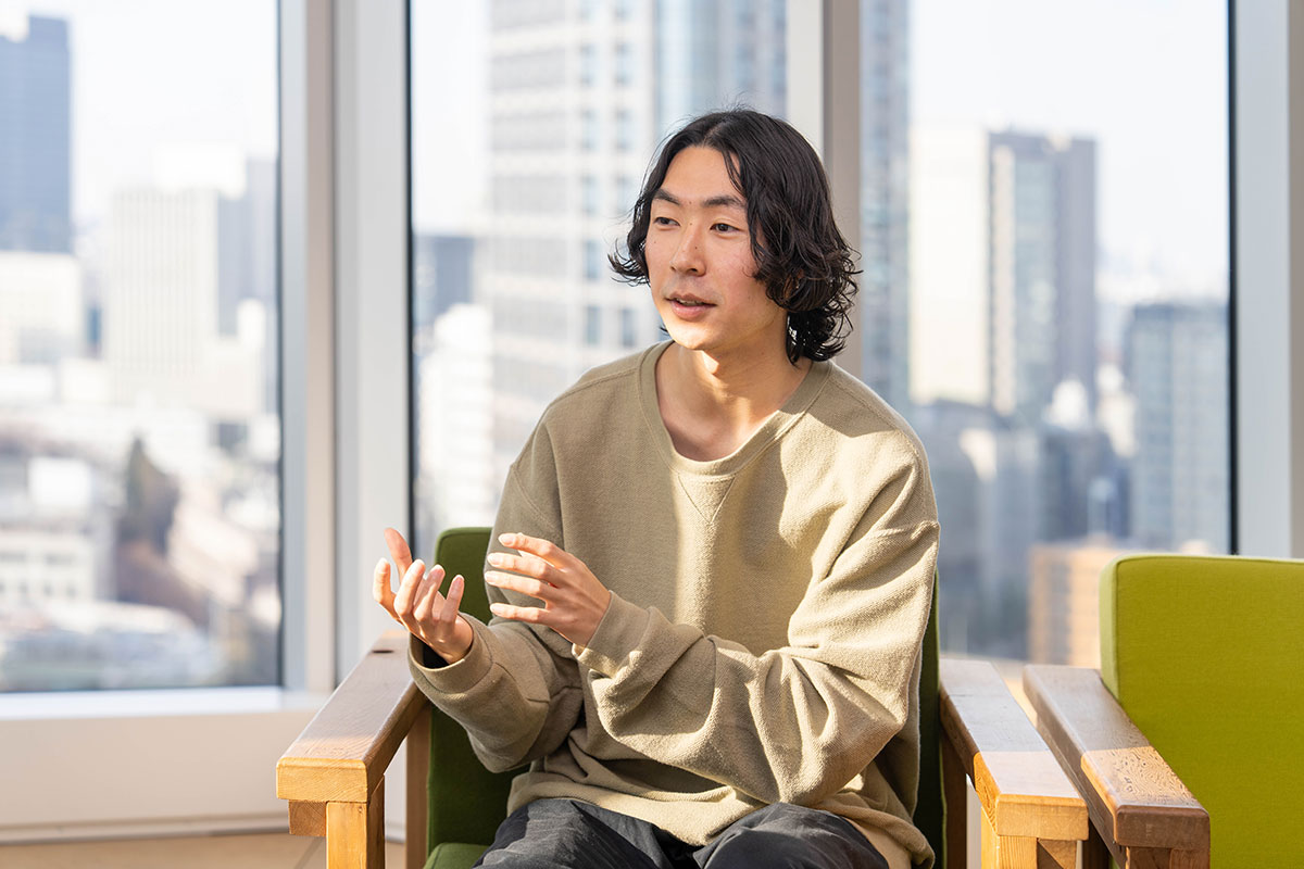 窓際の椅子に座る松浦の写真。肩までの黒髪を中央分けにし、ベージュのトップス姿で手振りを交えながら説明している。背後に都市の景色が広がる