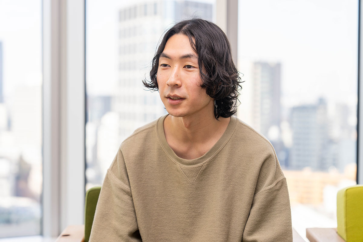 窓を背景に椅子へ座る松浦の上半身写真。中央分けの黒髪で、落ち着いた表情を浮かべながら前方を見つめている。柔らかな自然光が差し込んでいる