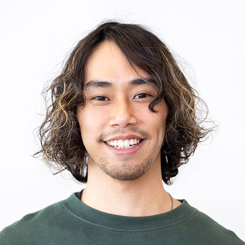 グレー背景の前で微笑む男性、百瀬のバストショット。肩にかかるゆるいパーマの黒髪にあごひげをたくわえ、緑のトップスを着用して歯を見せて笑っている。