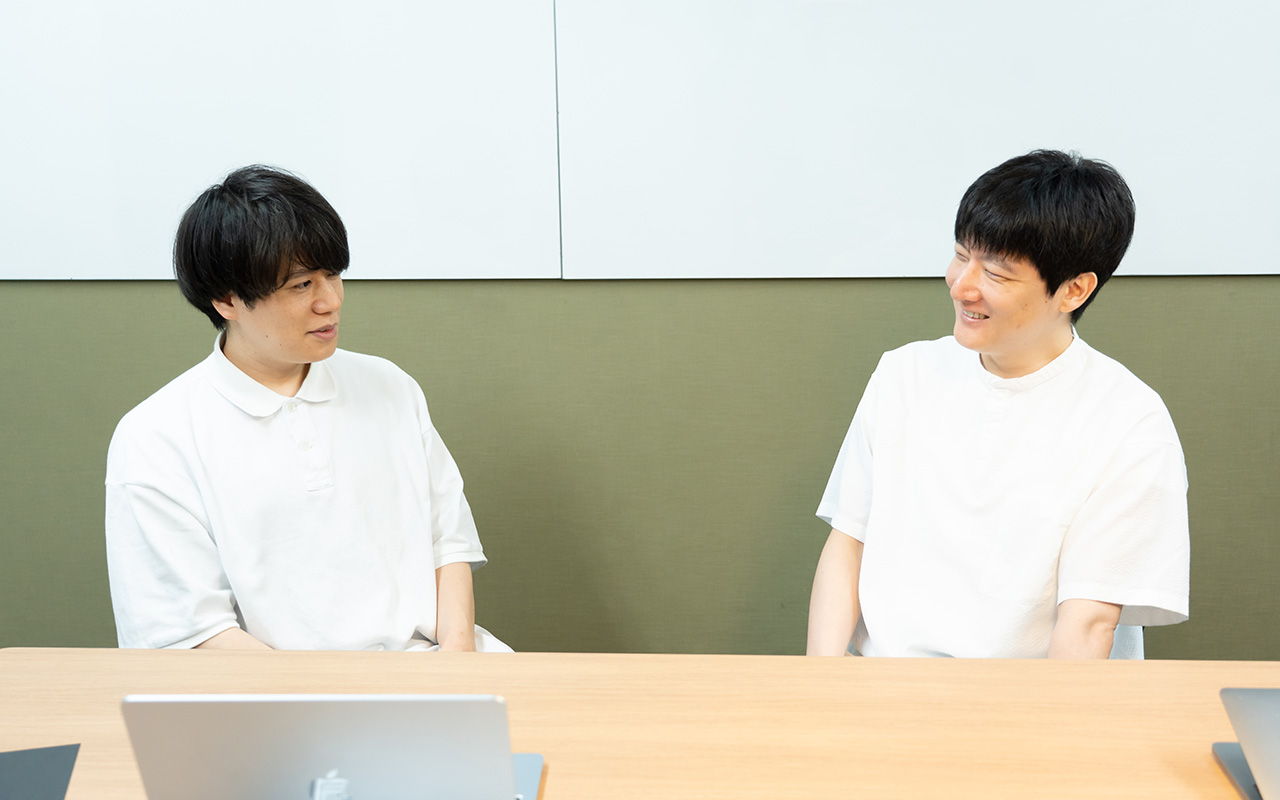 糠谷さんと石森さんが会話している様子。左側の糠谷さんは白いポロシャツを着用し、黒髪のミディアムヘアで、椅子に座って石森さんの方を向きながら落ち着いた表情で話している。右側の石森さんは白い半袖シャツを着用し、椅子に座って糠谷さんの方を見ながら笑顔を見せている。