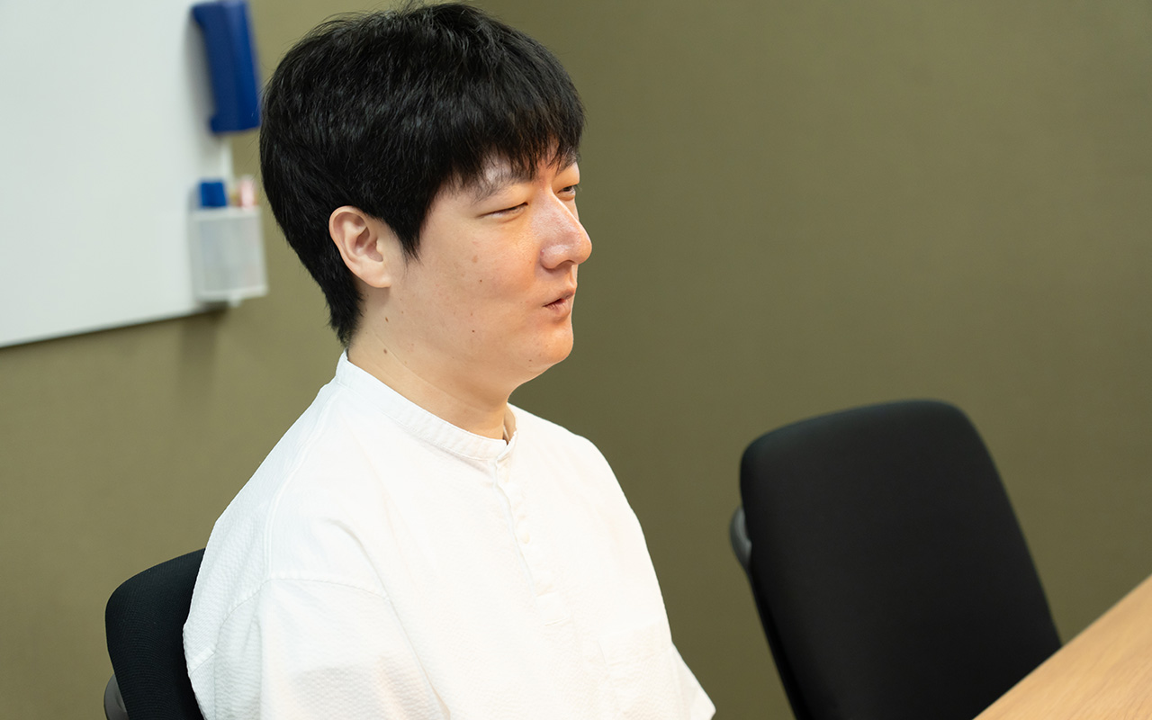 石森さんのバストショット。白い半袖シャツを着用し、短髪の石森さんが、椅子に座って横を向き、穏やかな表情で相手の話を聞いている様子