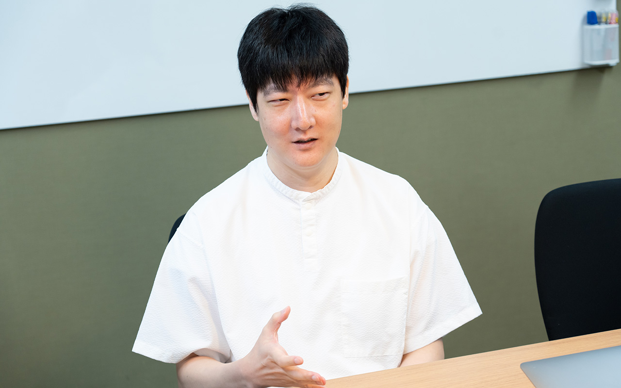 石森さんのバストショット。白い半袖シャツを着用し、短髪の石森さんが、椅子に座りながら片手を軽く前に出して身ぶりを交え、落ち着いた表情で話している様子