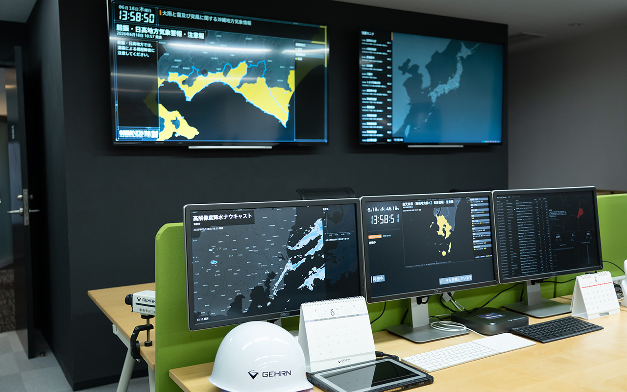 防災情報を監視・表示するオフィス内のモニタリングスペースである。壁面には大型ディスプレイが2台設置され、津波警報や地図情報、日本地図などの防災関連データが表示されている。デスク上には複数のモニターが並び、気象レーダーや津波予測、地図、一覧形式の監視情報などが映し出されている。