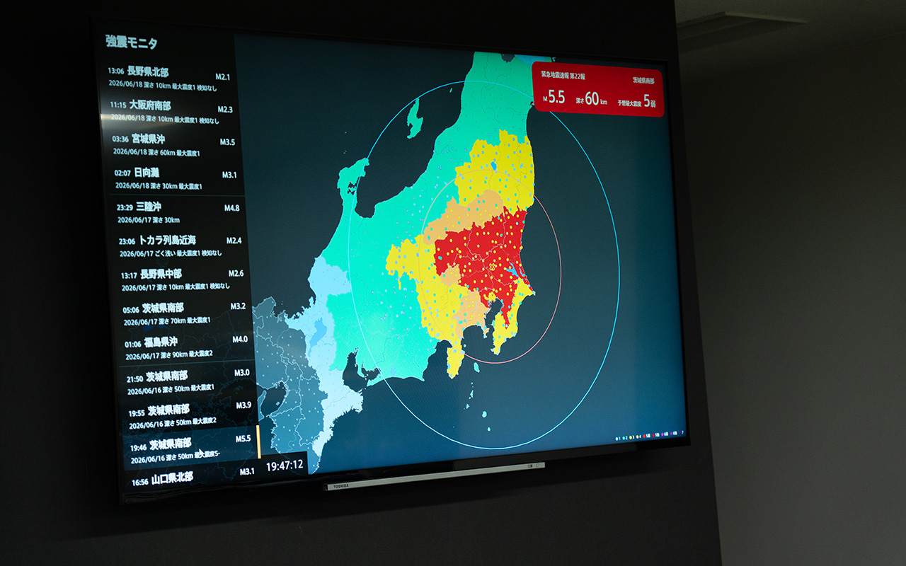 壁面に設置された大型モニターに、地震情報を表示している様子である。画面中央には関東地方を中心とした地図が表示され、震度分布が水色、黄色、橙色、赤色で色分けされている。