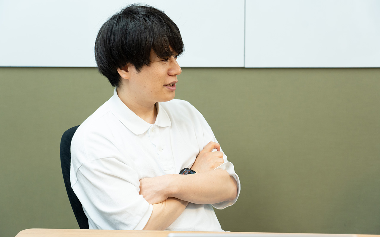 糠谷さんのバストショット。白いポロシャツを着用し、黒髪のミディアムヘアの糠谷さんが、椅子に座って腕を組みながら横を向き、真剣な表情で話している