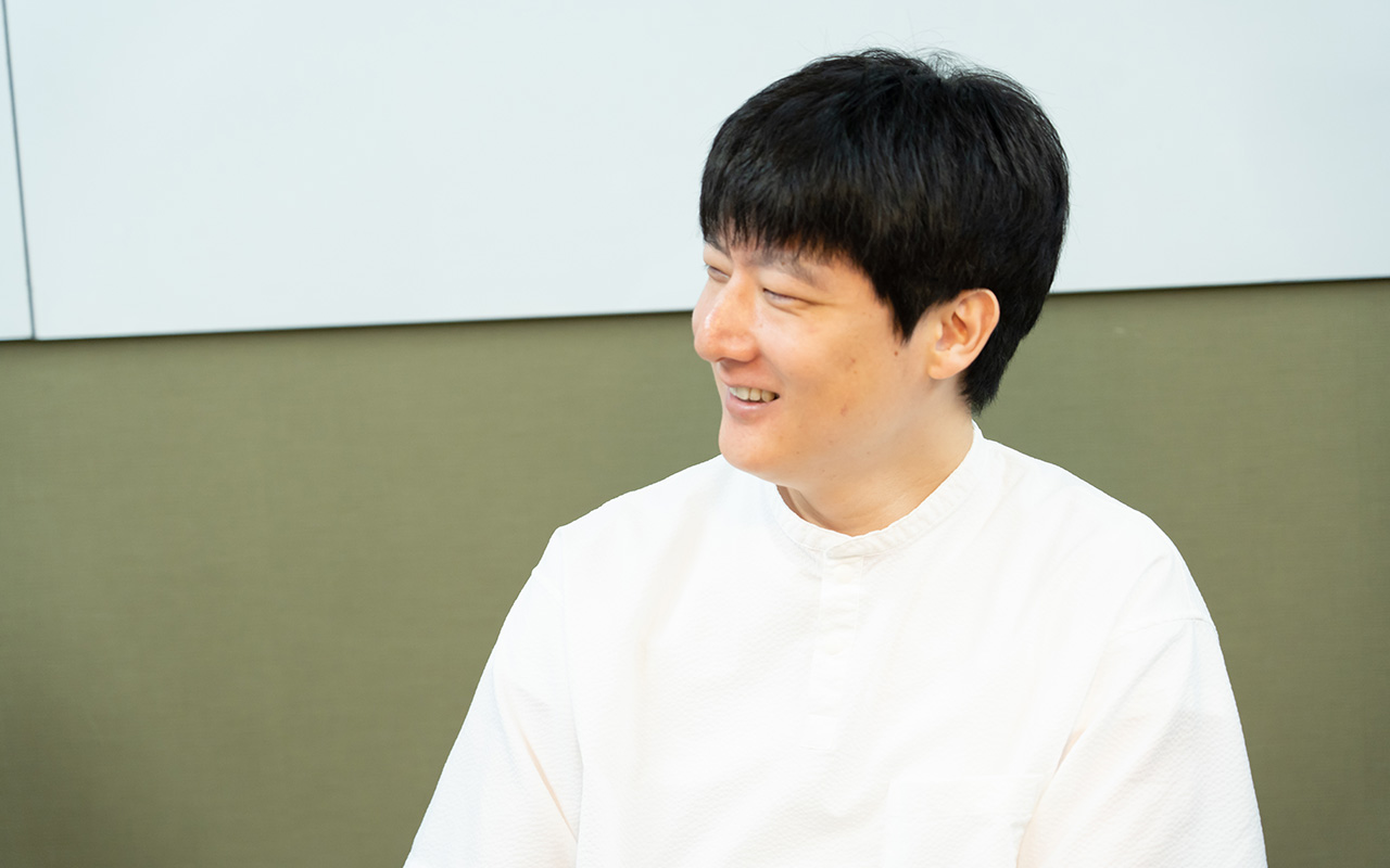 石森さんのバストショット。白い半袖シャツを着用し、短髪の石森さんが、椅子に座って体をやや横に向け、笑顔で相手の方を見ながら会話している様子