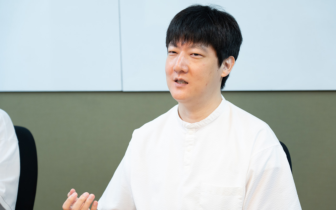 石森さんのバストショット。白い半袖シャツを着用し、短髪の石森さんが、椅子に座りながら片手を軽く上げて身ぶりを交え、落ち着いた表情で話している様子