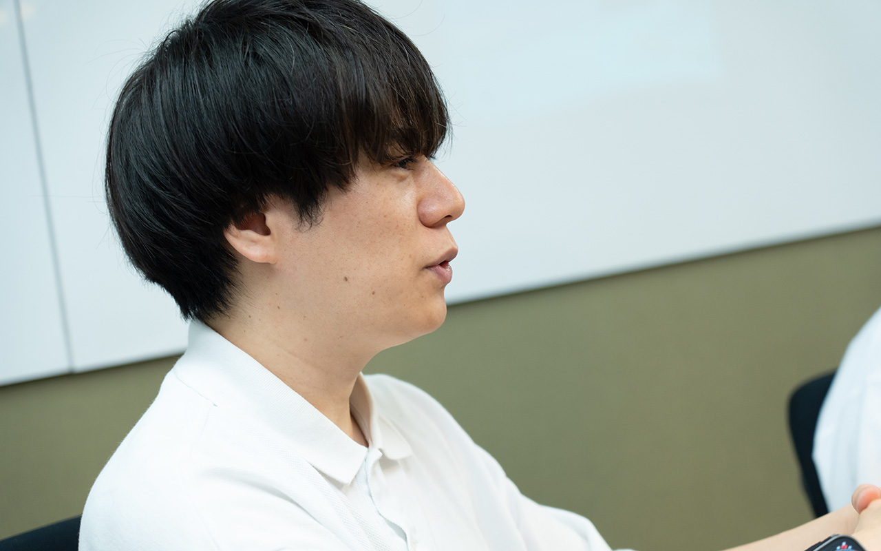糠谷さんの横顔のバストショット。白いポロシャツを着用し、黒髪のミディアムヘアの糠谷さんが、椅子に座って前方を向きながら、口を開いて相手に話しかけている様子