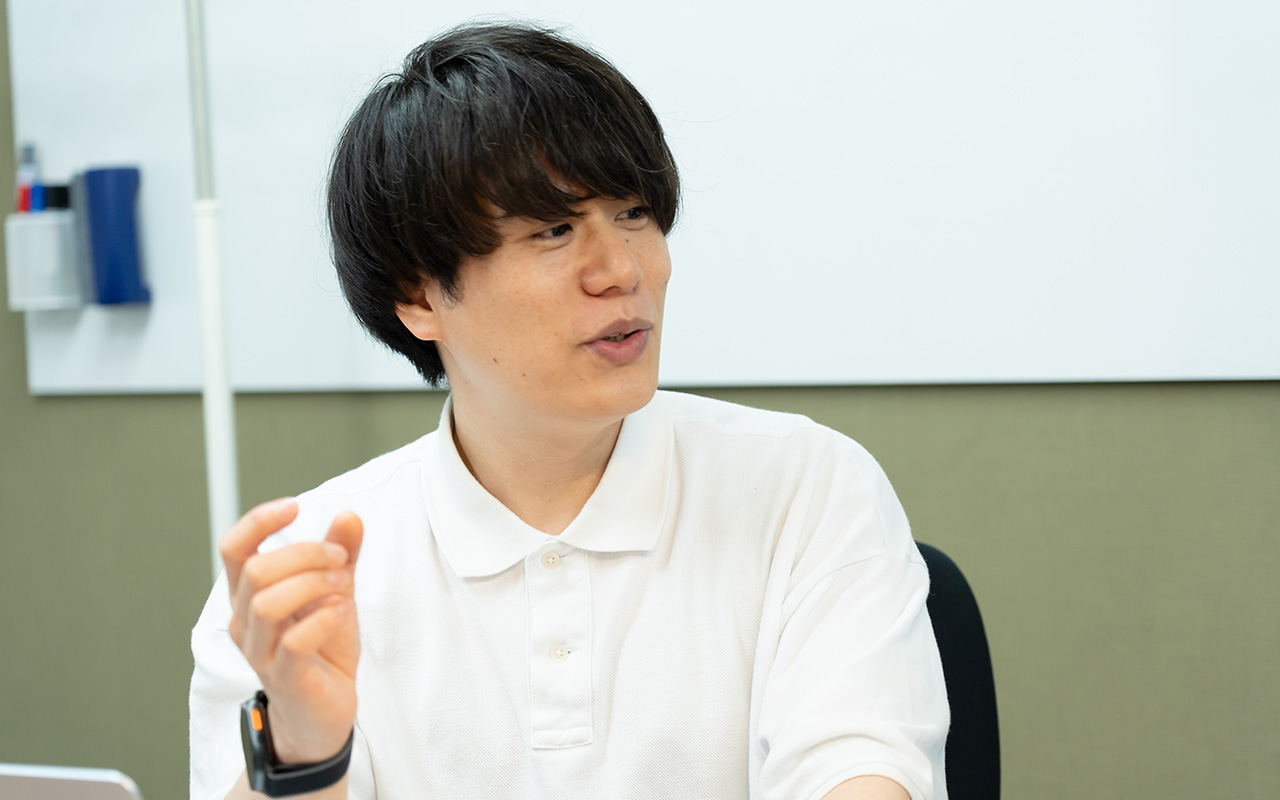 糠谷さんのバストショット。白いポロシャツを着用し、黒髪のミディアムヘアの糠谷さんが、椅子に座りながら片手を軽く上げて身ぶりを交え説明している様子