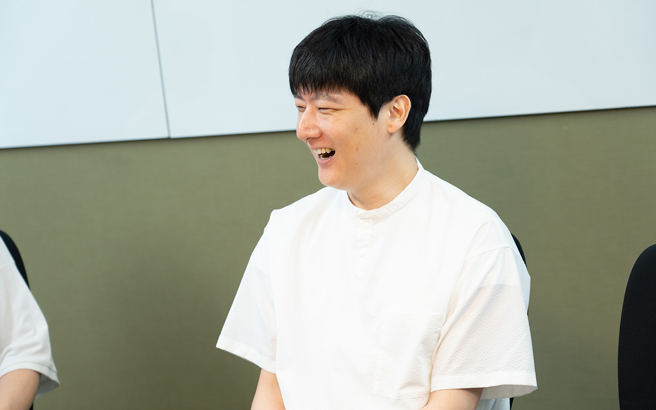 石森さんのバストショット。白い半袖シャツを着用し、短髪の石森さんが、椅子に座って顔を横に向け、大きく口を開けて笑っている様子