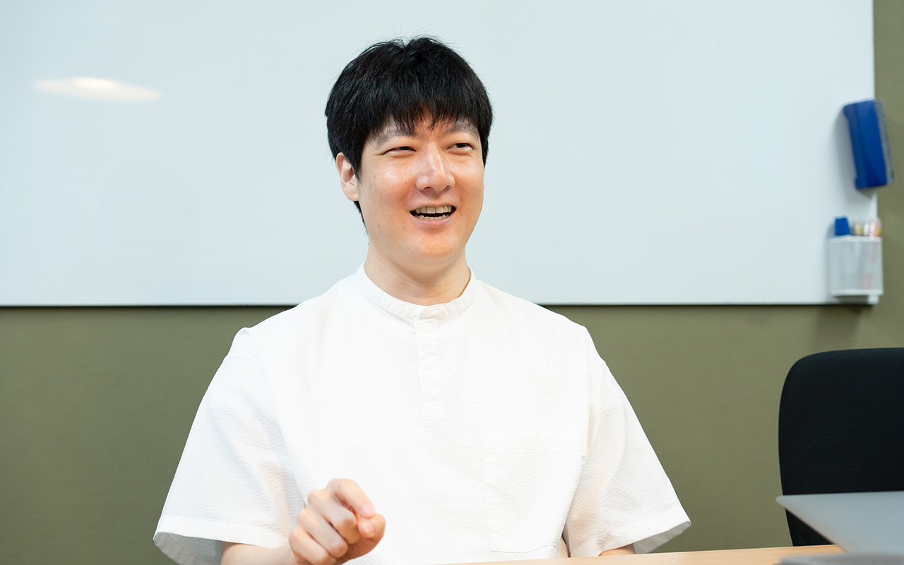 石森さんのバストショット。白い半袖シャツを着用し、短髪の石森さんが、椅子に座りながら人差し指を軽く前に向けて身ぶりを交え、笑顔で話している様子