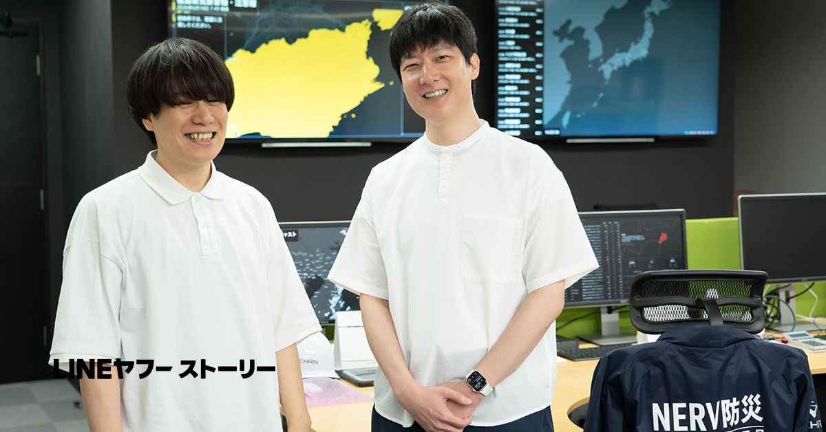 糠谷さんと石森さんの写真。背景は複数のモニターが設置されたオフィス空間で、大型モニターには日本地図やデータを表示した画面が映し出されている。デスクにはモニターやキーボードが並び、右手前の椅子には「NERV防災」と書かれたジャケットが掛けられている。