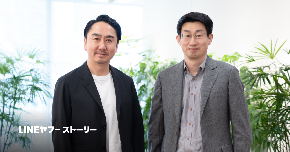 観葉植物のある室内で並んで立つ男性2人の写真。左は黒ジャケット、右はグレージャケット姿でカメラを見ている。左下に「LINEヤフーストーリー」と白文字がある