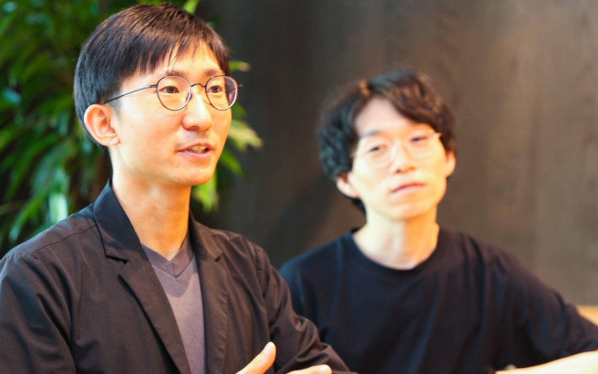 加藤のバストショット。グレーのVネックシャツの上に黒いジャケットを着ている。正面よりやや右方向を向き、話しながら手を前に出している