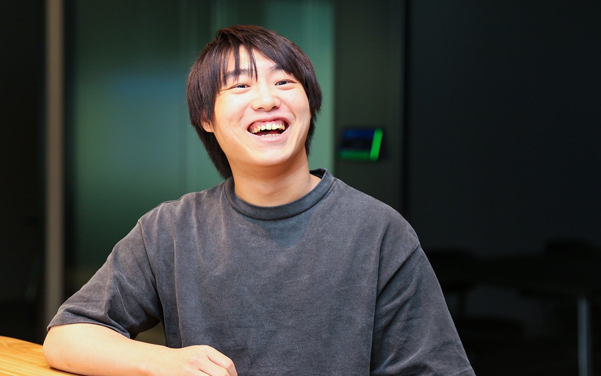 中山のバストショット。前ゆったりとした濃いグレーの半袖シャツを着ている。木製のテーブルに片腕を置き、正面よりやや右方向を向いて大きく笑っている
