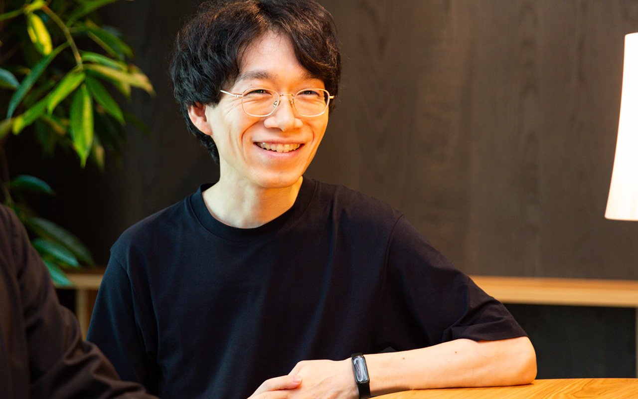 田中さんのバストショット。中央で分けた短い黒髪で細いフレームの眼鏡をかけ、黒い半袖シャツを着ている。木製のテーブルに腕を置き、正面よりやや左方向を向いて笑っている