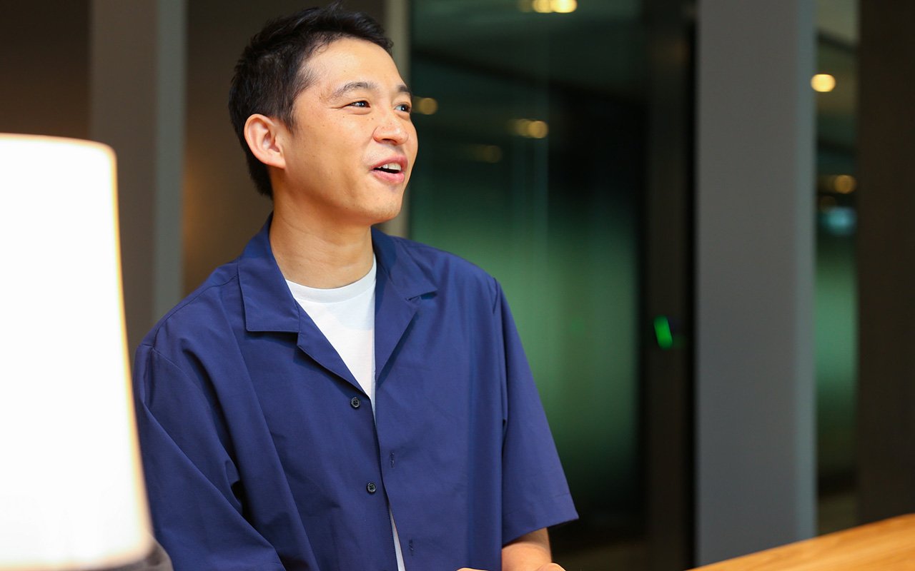 山田のバストショット。正面よりやや右方向に顔と視線を向け、笑顔で話している様子