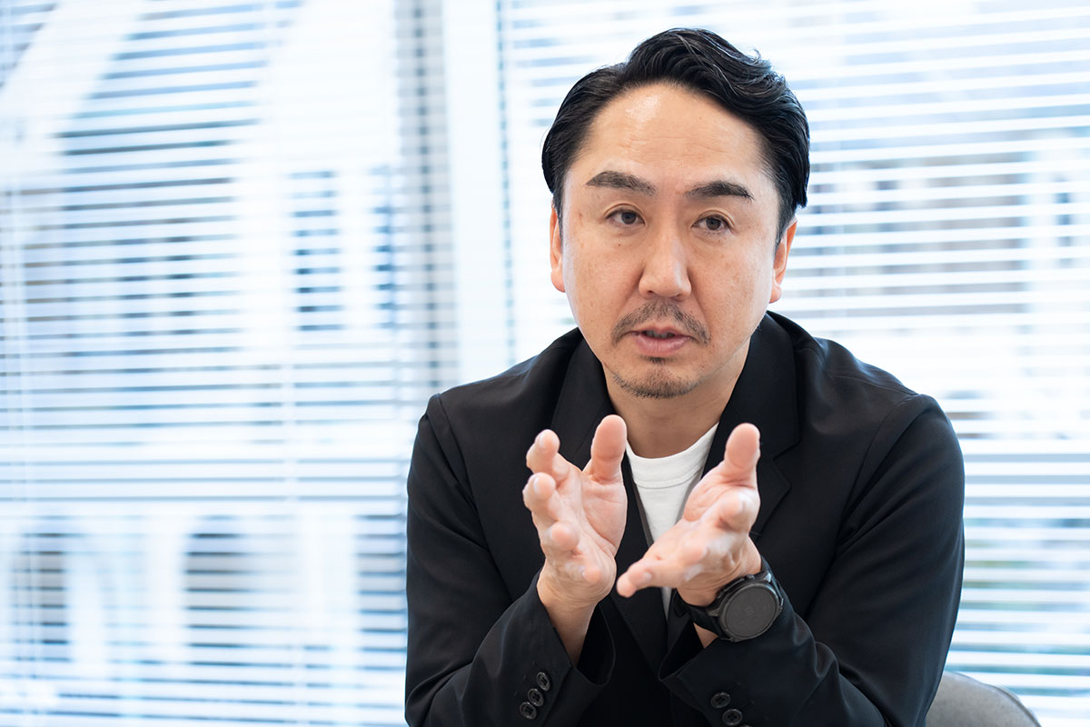 グレージャケットを着た慎の上半身写真。やや前傾姿勢で真剣な表情を見せ、前方を見つめながら話している。