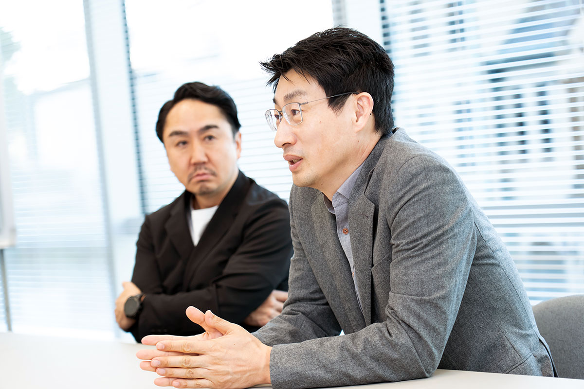 会議室で話す2人の写真。手前の慎はグレージャケットで手を組み説明し、奥の出澤はじっくりと話を聞いている。