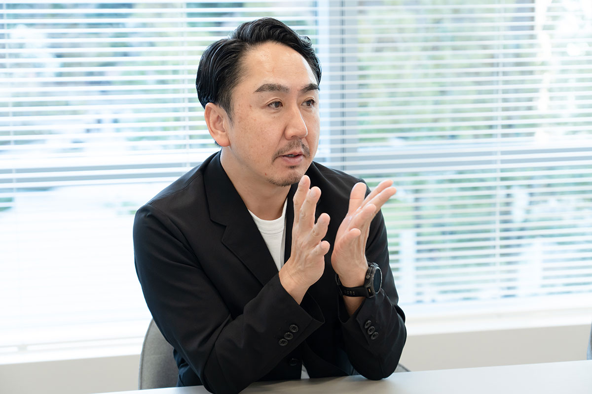 黒ジャケットの出澤が手を上げて説明する写真。真剣な表情で前方を見つめ、背景にはブラインド越しの光が差し込んでいる