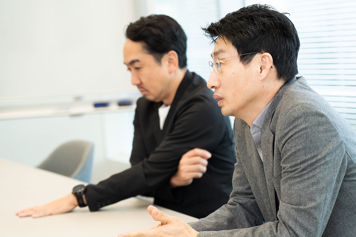 インタビューに応える2人の横からの写真。手前の慎はグレージャケットで話し、奥の出澤は黒ジャケットで机に手を置きながら聞いている。