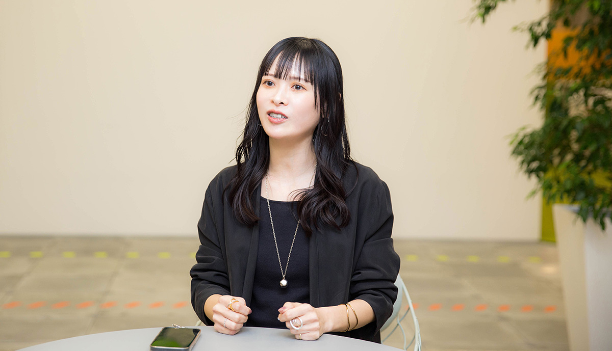 菅沼がオフィスのテーブルに座り、両手を軽く握りながら話している写真。