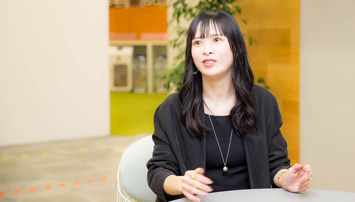 菅沼がオフィスのテーブルに座り、両手を軽く広げながら話している写真。