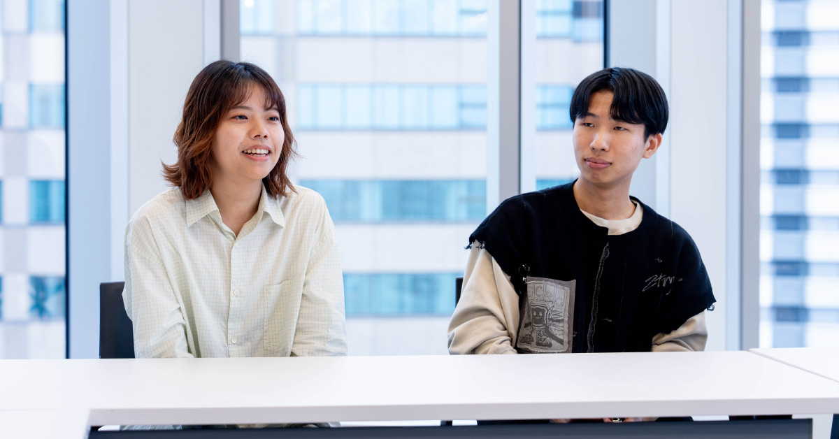 タスクフォースメンバー2人が並んで座り、笑顔を交えながら会話する写真。明るい窓際の会議室で撮影されている。