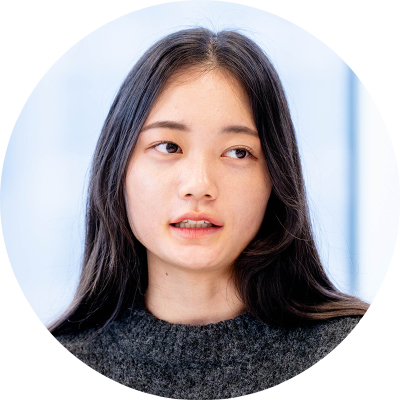 ダークグレーのセーターを着た、黒髪でロングヘアの女性の円形トリミング写真。視線を斜め左に向け、口を開いて何かを話しているような自然な表情を浮かべている。