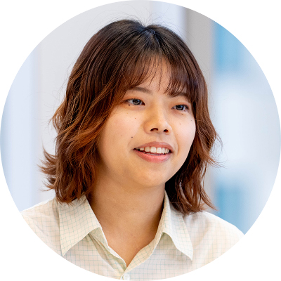 淡いグリーンのチェックシャツを着た、前髪のある茶色のミディアムヘアの女性の円形トリミング写真。視線を左方向に向け、穏やかな笑顔で話をしている様子。
