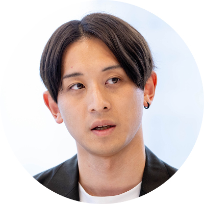 白いインナーに黒いジャケットを合わせ、左耳にフープピアスをしたセンター分けの短髪男性の円形トリミング写真。視線を左側に向け、落ち着いた表情で話をしている。