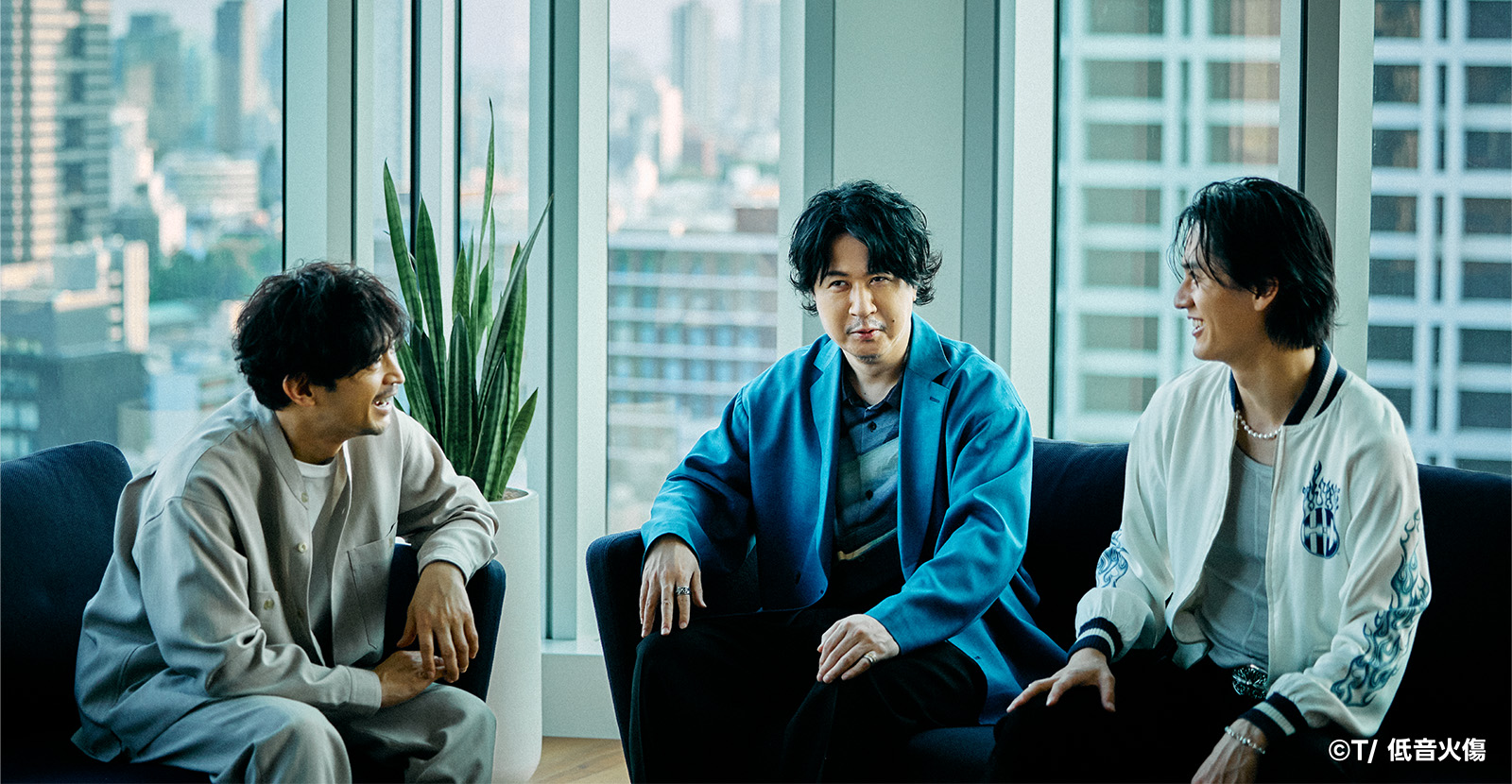 3人の男性がソファに座り、楽しそうに会話をしています。背景には大きな窓があり、都市の景色が広がっています。