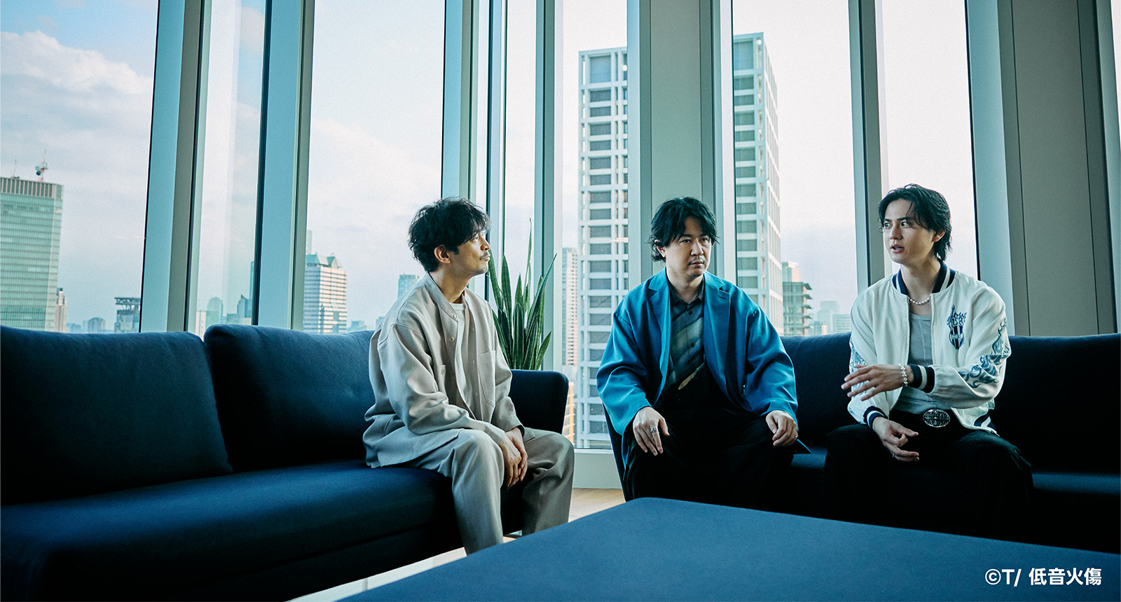  大きな窓のある部屋で3人の男性がソファに座り、話をしています。背景には都市の風景が見えます。