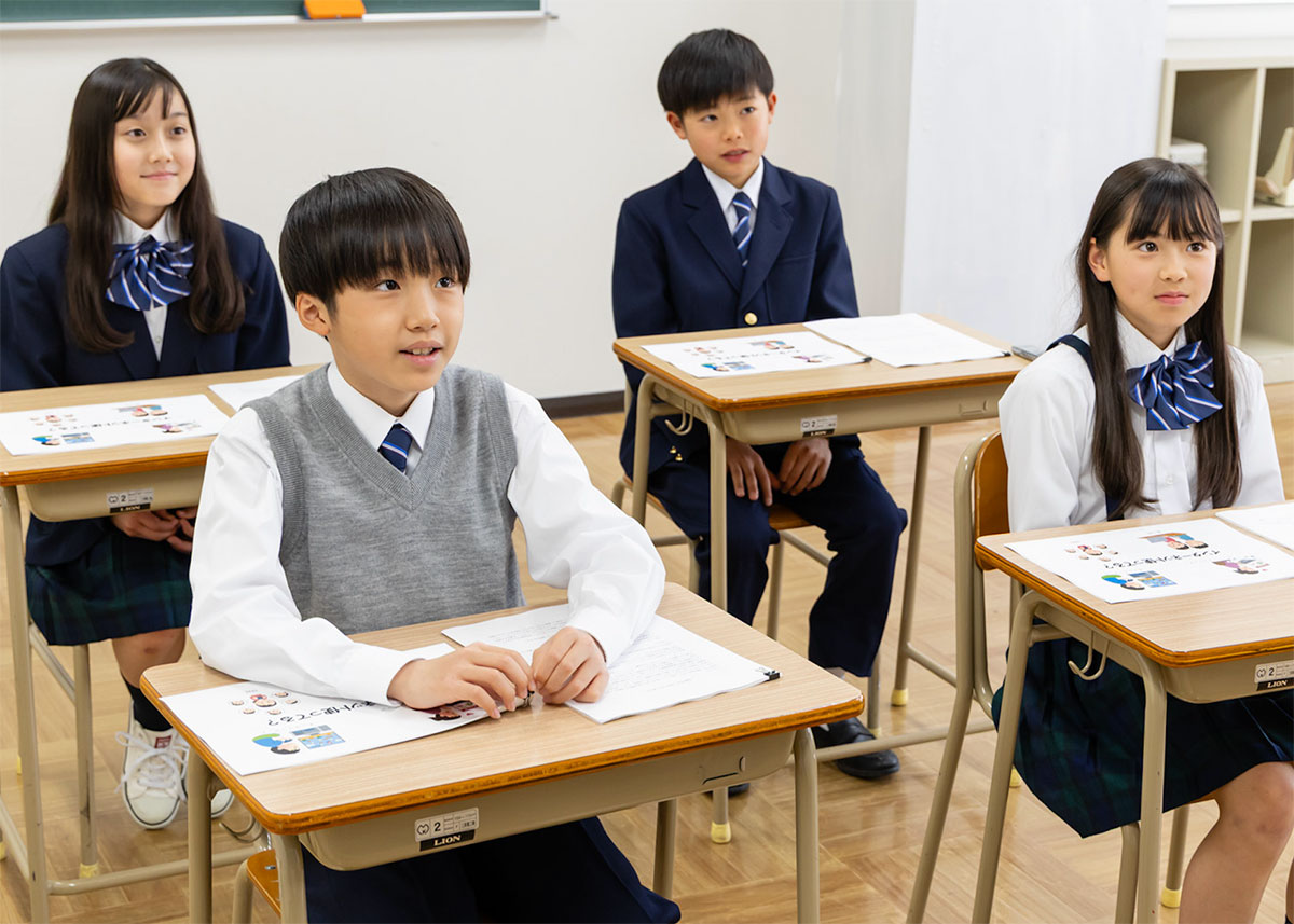 教室で制服を着た4人の児童・生徒が机に向かって座っている様子を写しています。前方の男の子が発言しており、周囲の3人は真剣に聞いている雰囲気です。学びの場での積極的な姿勢が伝わります