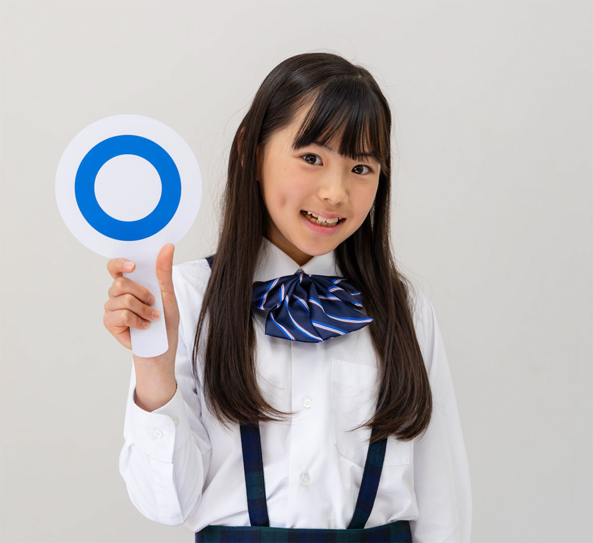 白いシャツとリボンを着た少女が、青い丸印の札を持ってほほ笑んでいる様子です