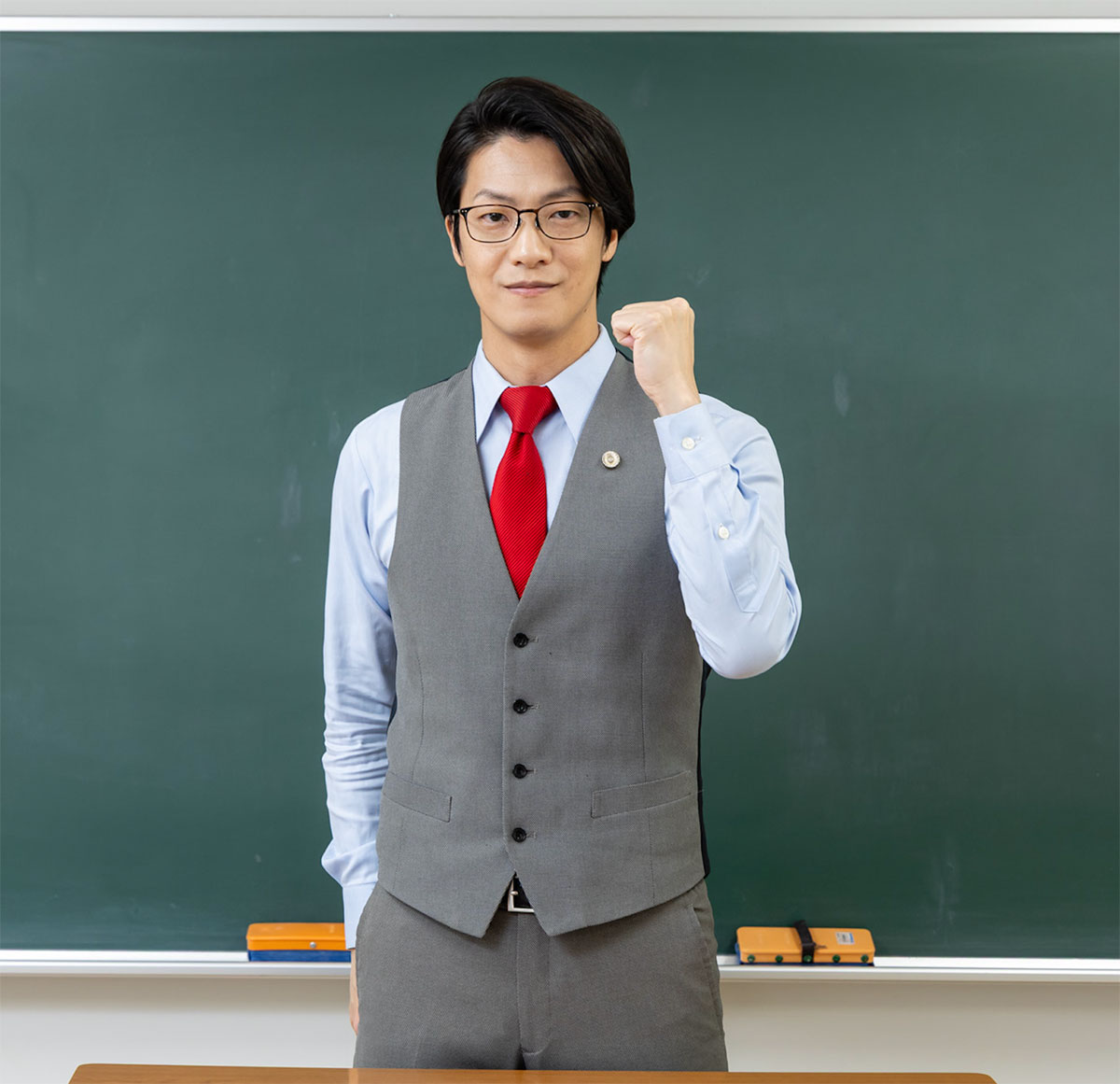 こたけ正義感さんが黒板の前でガッツポーズをしている様子です。彼はグレーのベストと赤いネクタイを着用しています