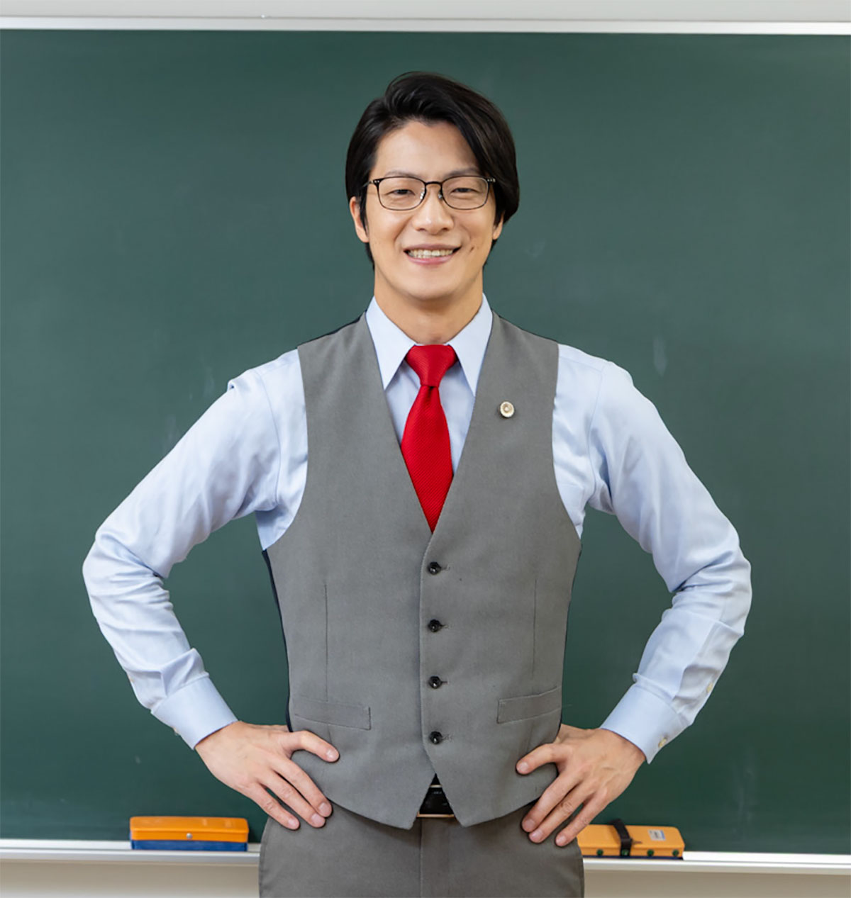 黒板の前に立つスーツ姿の男性が微笑んでいる様子です。彼は手を腰に当てて自信に満ちた表情をしています