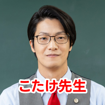 眼鏡をかけた男性・こたけ正義感さんが笑顔で写っているクローズアップ写真です。彼はグレーのベストと赤いネクタイを着用しています