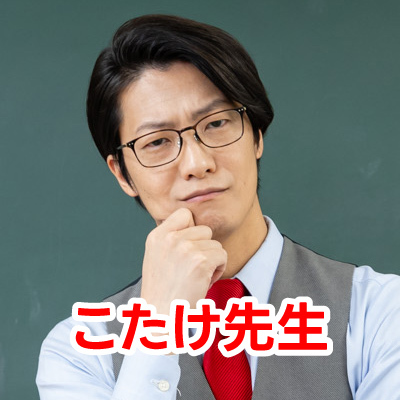 眼鏡をかけた男性・こたけ正義感さんが笑顔で写っているクローズアップ写真です。彼はグレーのベストと赤いネクタイを着用しています