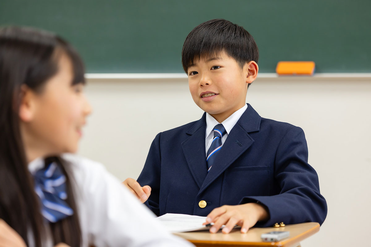 男の子が教室で笑顔で話している場面です。隣の席の女の子と楽しそうに会話をしています