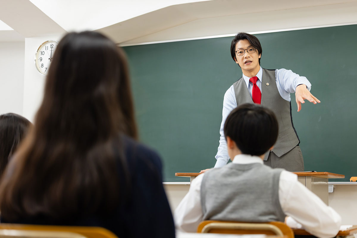 教師が生徒に何かを説明している場面です。彼は手を使いながら熱心に指導しています