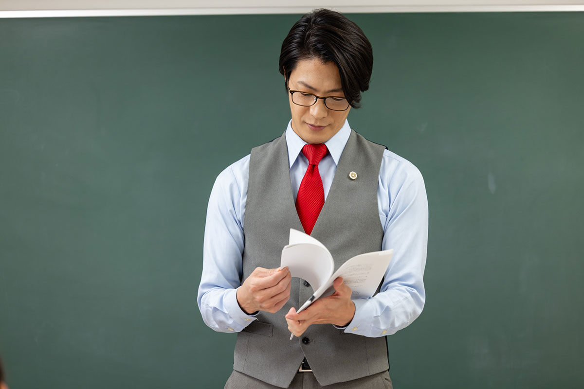 教師が教科書を見ながら授業を進めている様子を示しています。彼は黒板の前で資料を確認しています